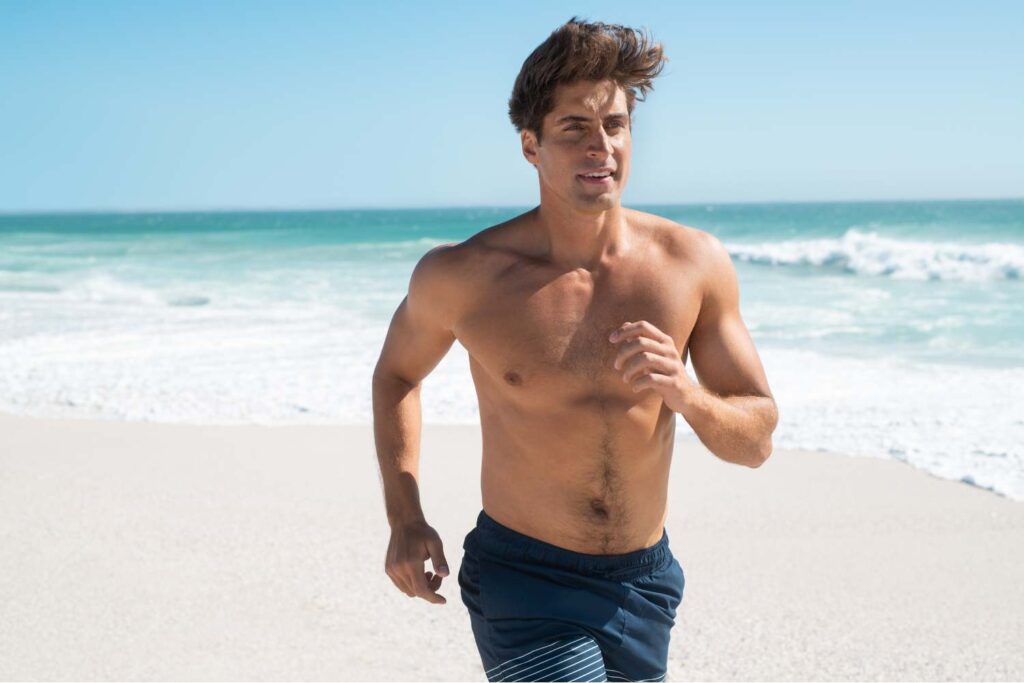 Shirtless man in navy swim shorts jogging along a sandy beach with ocean waves behind him